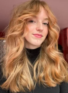 Vrouw met halflang, golvend haar in een warme blonde kleur met koperachtige toner, draagt een zwarte coltrui.
