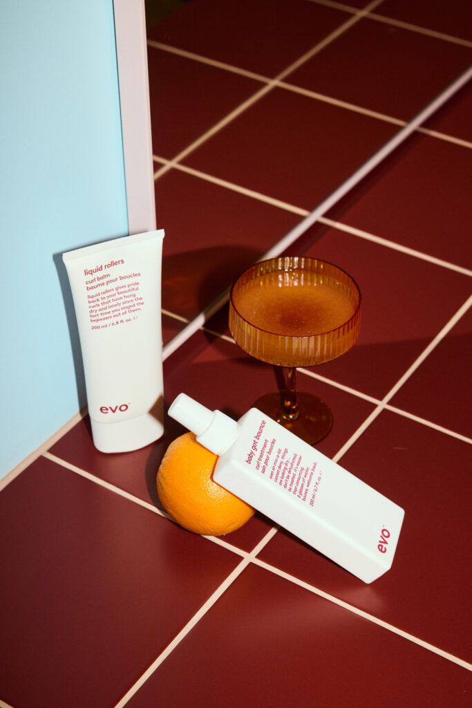 Twee evo-haarproducten staan gestileerd op een rood betegelde ondergrond naast een sinaasappel en een amberkleurig glas.