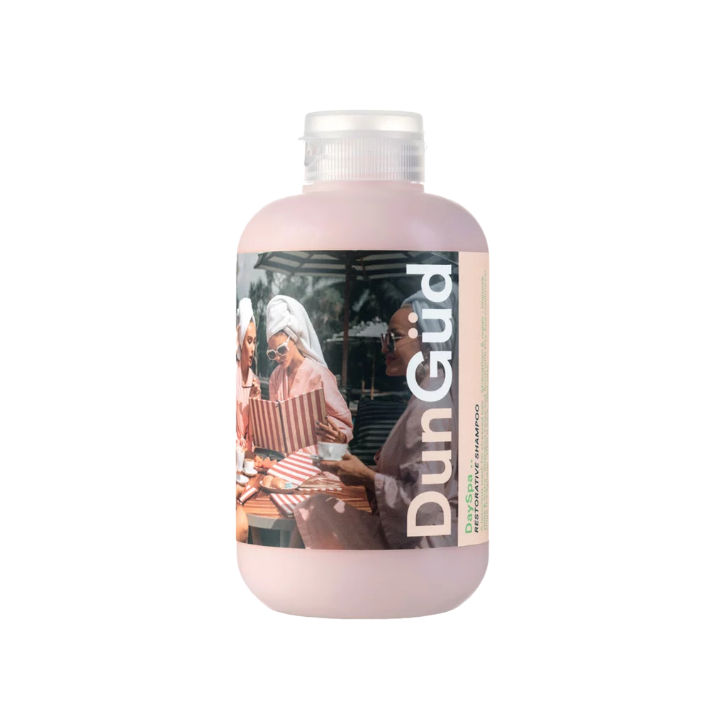 Roze fles DunGüd Day Spa Restorative Shampoo met een etiket waarop vrouwen in badjassen ontspannen bij een spa.