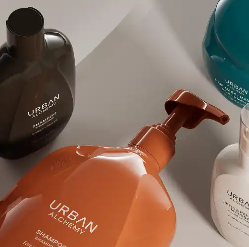 Vier flessen Urban Alchemy haarproducten in verschillende kleuren en formaten liggen op een lichtgrijze ondergrond.